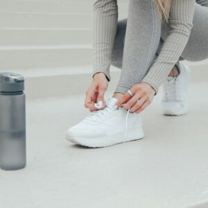 Young woman tying white sports shoe. Grey sport bottle with water